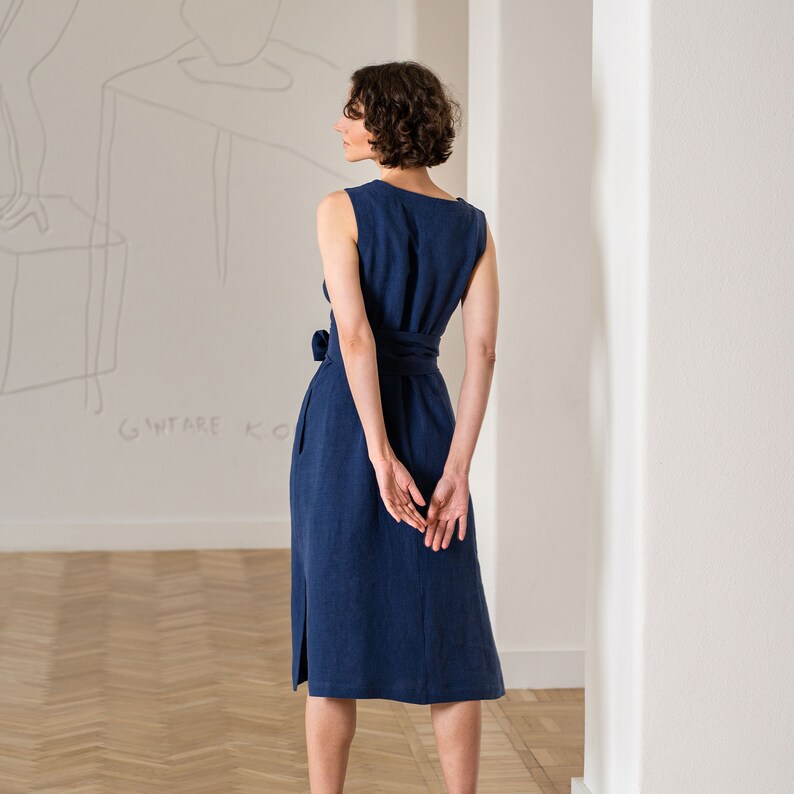 Photograph features a woman wearing the HAILEY summer wrap dress in storm blue color. The woman is visible from the back. Her hands are behind her back. The posture of the model help reveal the side, where the grafts are at the bottom of the dress.