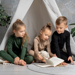 Puede incluir: Tres niños con camisetas y pantalones de manga larga a juego leen un libro. Uno viste de verde, otro de color beige y el tercero de negro. Están sentados en una alfombra gris frente a un tipi blanco.