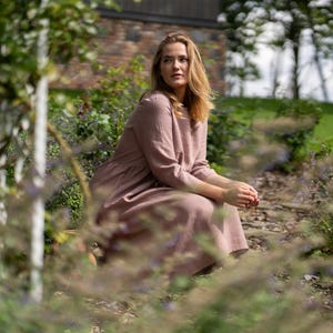 May include: A dusty rose-colored midi dress with long sleeves, a round neckline, and a gathered waistline. The garment features a simple, solid-colored design with a visible texture, falling to a mid-calf length with a relaxed, loose-fitting silhouette.