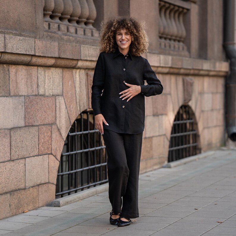 May include: A woman wearing a black linen shirt and matching pants. She is standing in front of a brick building and smiling.