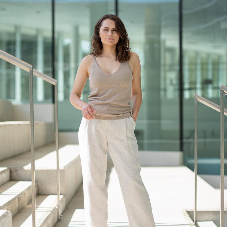 A woman with shoulder-length wavy brown hair stands looking directly at the camera, with one hand in her pocket. She wears menique tank top and wide-legged white pants Lotus, positioned on light-colored stairs with silver handrails. A modern building is in the background.