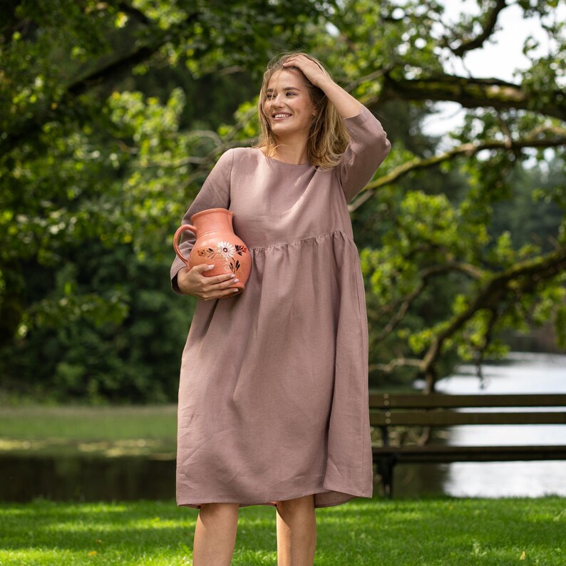 May include: A dusty rose-colored knee-length dress with long sleeves, a round neckline, and a loose, flowing silhouette. The garment is held alongside a terracotta-colored ceramic pitcher featuring a hand-painted floral design in white and green.