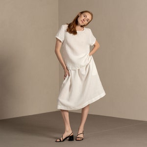 The model is wearing the SOPHIA skirt in pure white. She has one hand inside the side pocket, and the other on her side. She has one of her feet put to the side, tilting the hip and her head. The skirt has deep pockets, as can be seen in the picture.