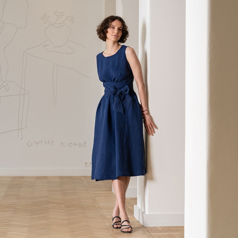 Photograph features a woman wearing the HAILEY summer wrap dress in storm blue color. The dress design is suitable for all shapes and sizes, because of the wrap design and included obi belt. The design is sleeveless, with a round neckline.