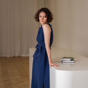Photograph features a woman wearing the HAILEY summer wrap dress in storm blue color. The woman is leaning on a tabletop. The sideview of the picture accentuates the waist.