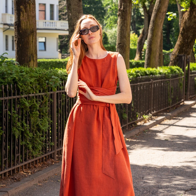 May include: A woman wearing a rust-colored linen dress with a self-tie belt. She is wearing sunglasses and has red hair.