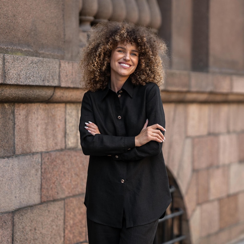 May include: A black linen shirt with a button-down collar and long sleeves. The shirt is worn by a person with curly hair and a smile.