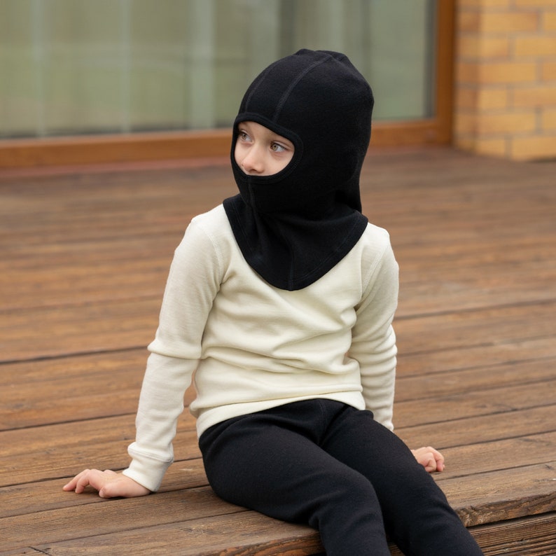 May include: A child wearing a black balaclava and a white long-sleeved shirt with black pants. The child is sitting on a wooden surface.