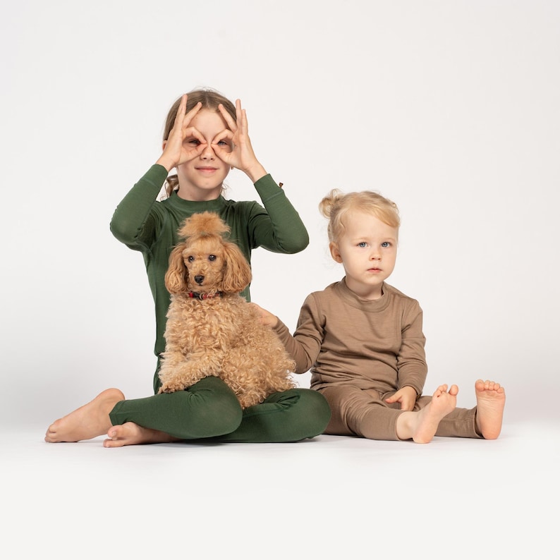 Two children seated on the floor; an older child in green holds a small apricot-colored poodle while a younger child in light brown sits beside them.