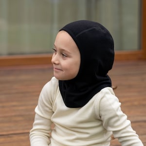 May include: A young person wearing a black fleece balaclava and a white long-sleeved shirt. The person is sitting on a wooden floor.
