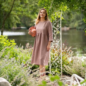 May include: A dusty rose-colored midi dress with long sleeves, a round neckline, and a gathered waistline. The dress is made of a solid-colored fabric and is held alongside a terracotta-colored ceramic pitcher with a handle and a spout.