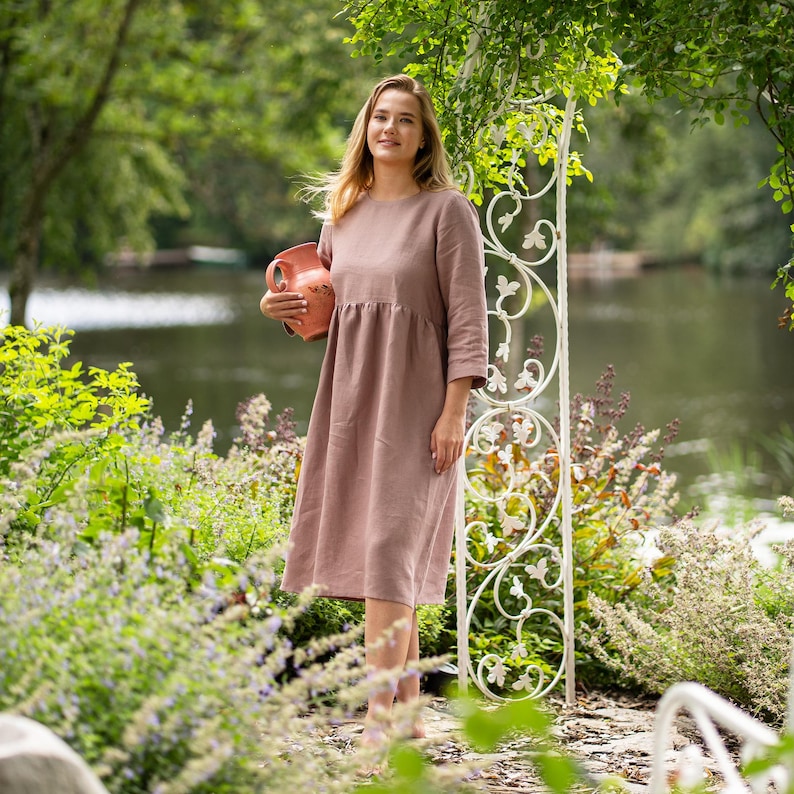 May include: A dusty rose-colored midi dress with long sleeves, a round neckline, and a gathered waistline. The dress is made of a solid-colored fabric and is held alongside a terracotta-colored ceramic pitcher with a handle and a spout.