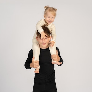 May include: A long-sleeved cream-colored shirt and matching pants set worn by a small child, and a long-sleeved black shirt and matching pants set worn by an older child. Both sets feature a simple, solid-colored design with ribbed cuffs.