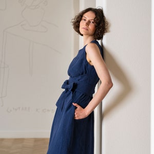 Photograph features a woman wearing the HAILEY summer wrap dress in storm blue color. The woman is leaning on a wall in a room. The side view features the sleeveless design and side pockets. The model’s hand is in one of them.