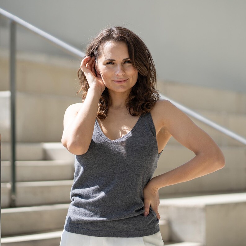 A woman with wavy brown hair looks to her right, hand on hip, wearing a dark gray tank top and white menique trousers. She stands on light-colored stairs with silver railings.
