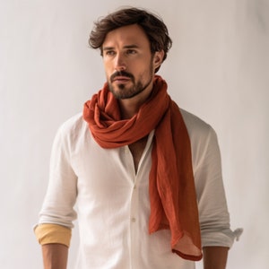 May include: A man wearing a white button-down shirt with a long, rust-colored scarf tied around his neck. The scarf is draped loosely around his neck and hangs down to his chest.