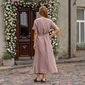 The model wears the ELEANOR maxi wrap dress in faded rose. She has one hand on her hips, while the other one hangs loosely. She has her back turned to the camera, the waist is framed with the belt. The dress features a loose fit with a flowy skirt.