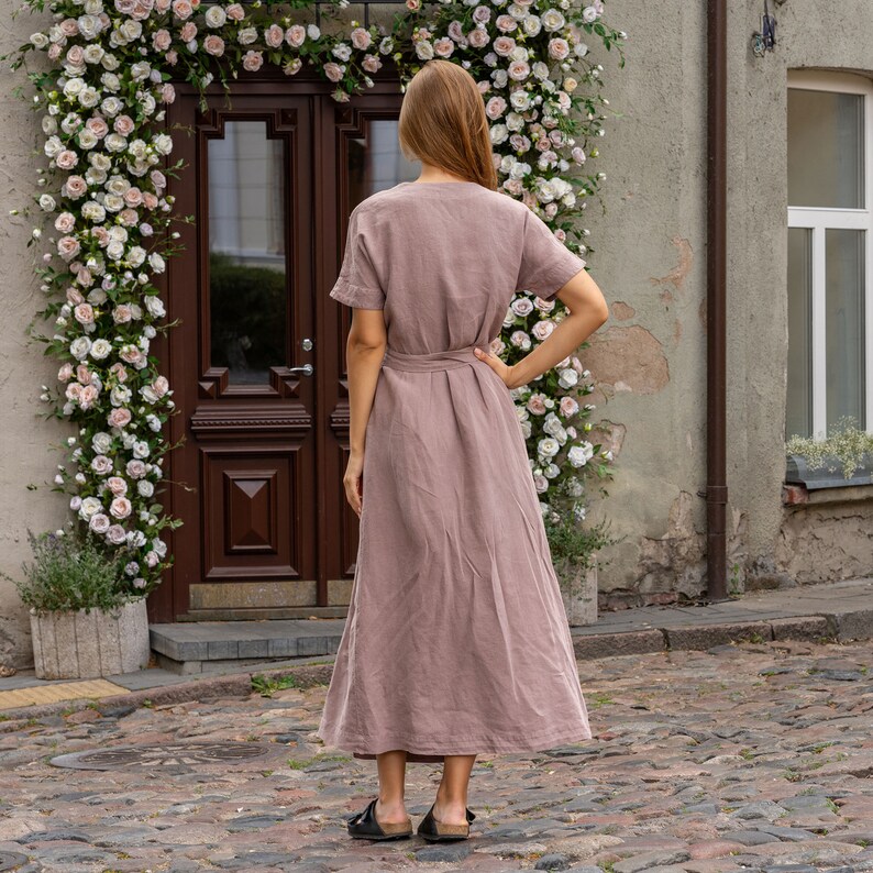 The model wears the ELEANOR maxi wrap dress in faded rose. She has one hand on her hips, while the other one hangs loosely. She has her back turned to the camera, the waist is framed with the belt. The dress features a loose fit with a flowy skirt.