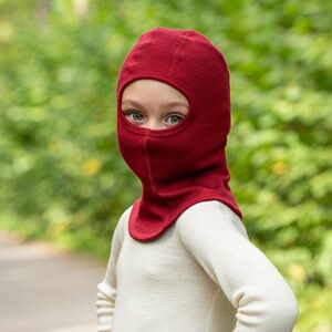 May include: A child wearing a red balaclava made of wool. The balaclava covers the child's head and neck, leaving only their eyes visible.