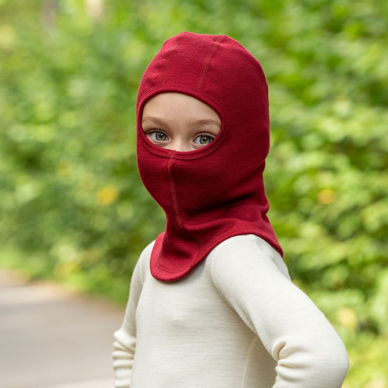 May include: A child wearing a red balaclava made of wool. The balaclava covers the child's head and neck, leaving only their eyes visible.