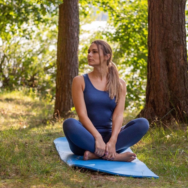 K&ouml;nnte beinhalten: Eine Frau sitzt im Schneidersitz auf einer blauen Yogamatte in einer Waldlandschaft. Sie tr&auml;gt ein blaues Tanktop und eine blaue Yogahose.