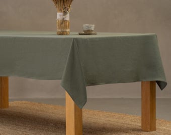 May include: A green linen tablecloth draped over a wooden table with a natural fiber rug underneath. The tablecloth has a simple, elegant design and is perfect for a casual dining setting.