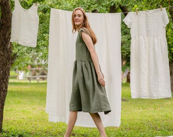 May include: A woman wearing a green linen dress walks in a grassy area with white linen clothing hanging on a clothesline behind her.