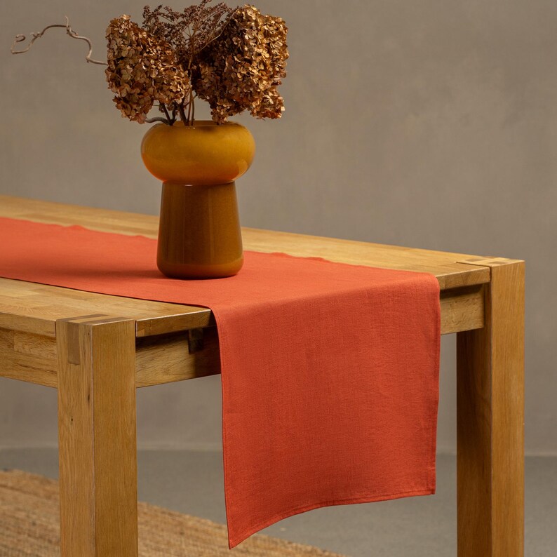 The menique cinnamon red color linen table runner on top of a wooden table. There is a vase on top of the runner.
