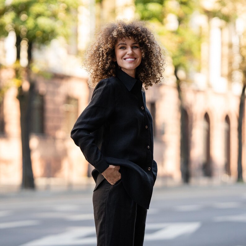 May include: A woman with curly brown hair is wearing a black long-sleeved shirt with a collar. She is smiling and has her hand in her pocket.