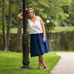 A woman rests casually against an ornate black lamppost in a lush green park with a tranquil lake in the background. She models a sleeveless pure white linen top and a storm blue midi skirt, completing the relaxed outfit with brown sandals.