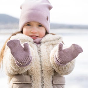 以下が含まれることがあります： ピンクのニット帽とふわふわのクリーム色のジャケットを着た少女。ピンクのニットの手袋をしています。