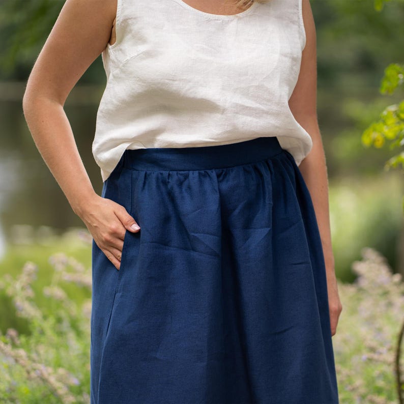 A close-up shot focuses on a woman's midsection as she stands outdoors, resting one hand comfortably inside the pocket of her skirt. The outfit features a relaxed-fit, pure white sleeveless linen top paired with a gathered storm blue midi skirt.