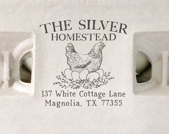 Op de afbeelding: Een wit kartonnen eierdoosje met een bruine rubberen stempel waarop "THE SILVER HOMESTEAD", een tekening van een hen en het adres "137 White Cottage Lane Magnolia, TX 77355" staat.