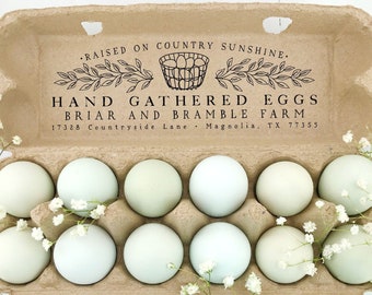 Op de afbeelding: Een dozijn lichtgroene eieren in een bruine kartonnen eierdoos. De doos is bedrukt met de tekst "RAISED ON COUNTRY SUNSHINE. HAND GATHERED EGGS BRIAR AND BRAMBLE FARM". Witte bloemen zijn rond de eieren gerangschikt.