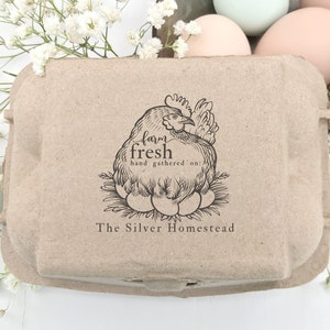 May include: A brown cardboard egg carton with a black and white illustration of a hen sitting on a nest of eggs. The text "farm fresh hand gathered" and "The Silver Homestead" is printed on the carton.