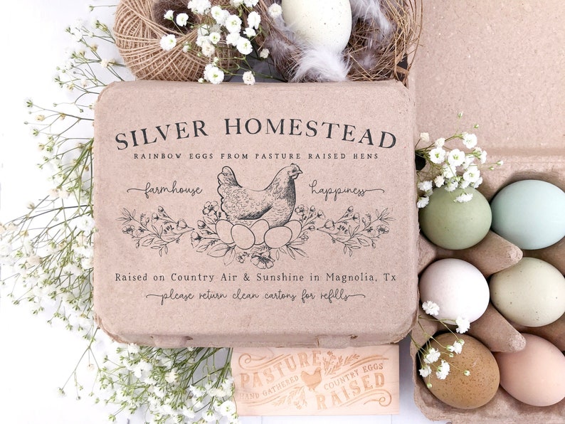 以下が含まれることがあります： Silver Homesteadの虹色の卵が入ったカートン。テキストと鶏のイラストが描かれています。カートンは白い花に囲まれ、様々な色の卵が入っています。「Pasture Raised」と書かれた小さな看板があります。
