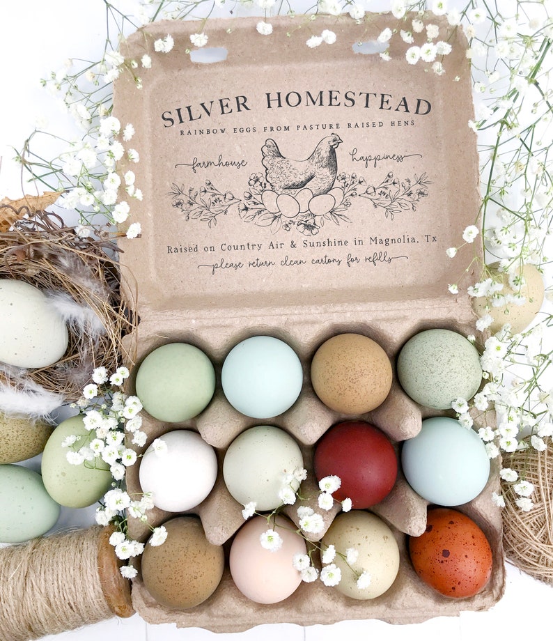 以下が含まれることがあります： 「SILVER HOMESTEAD」と「RAINBOW EGGS FROM PASTURE RAISED HENS」の文字が入った、段ボール箱に入った12個のレインボーカラーの卵。卵は白い花と羽の付いた巣に囲まれています。箱には「farmhouse」と「happiness」と書かれています。