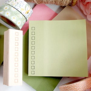 May include: A light green sticky note with a checklist, a wooden stamp, pink pen, and decorative washi tape rolls with floral and polka dot patterns. The items are arranged on a background of pink, green, and brown paper, with pink flowers.