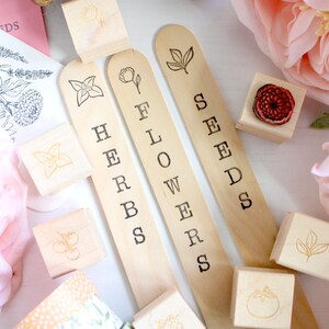 May include: Wooden garden markers labeled "HERBS", "FLOWERS", and "SEEDS" with stamped botanical illustrations. Accompanying wooden stamps feature various plant designs. Soft pink flowers and botanical drawings are in the background.