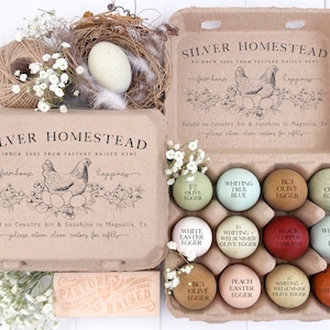 以下が含まれることがあります： 「Silver Homestead」と「Rainbow Eggs From Pasture Raised Hens」の文字が入った段ボール製の卵パック。パックには、それぞれに名前が付けられた、色とりどりの卵が1ダース入っています。卵と花が入った巣も写っています。