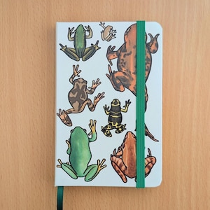 Peut inclure: Un petit carnet blanc orné d'illustrations colorées de différentes grenouilles. La couverture présente des grenouilles vertes, marron et orange. Il est doté d'un élastique vert et d'un marque-page en ruban noir.
