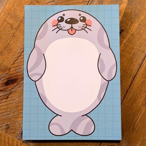May include: A rectangular notepad with a cartoon seal illustration. The seal is light grey with darker grey stripes, a white belly, and a pink tongue. The background is a light blue grid pattern.