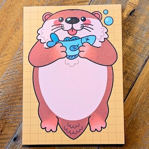 May include: A notepad featuring a cartoon otter holding a blue fish. The otter is brown and pink with a white ruff and a happy expression. The background is a yellow grid.
