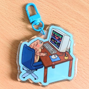 May include: A clear acrylic keychain featuring a cartoon frog at a computer desk. The frog is brown and tan, sitting in a blue chair, and working on a retro computer. The keychain has a blue clasp.