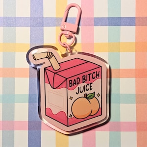 Könnte beinhalten: Ein rosafarbener Acryl-Schlüsselanhänger mit einem Saftbox-Design. Der Text "BAD BITCH JUICE" und eine Pfirsich-Illustration sind zu sehen. Er hat einen rosafarbenen Verschluss und Ring. Der Hintergrund ist ein farbenfrohes Karomuster.