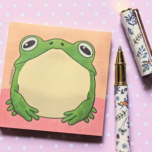 May include: A green frog with a light brown background. The frog is sitting on a pink sticky note with the text "Remember" written on it.