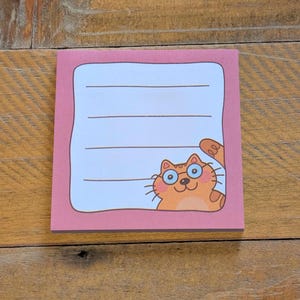 May include: A square pink sticky note pad with a light blue interior and lined pages. A cartoon orange cat wearing glasses peeks from the corner. The notepad is on a wooden surface.