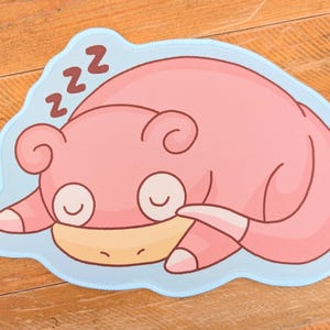 May include: A pink cartoon Slowpoke character is shown sleeping on a light blue mat with a wavy edge. The character has closed eyes and the word "zzz" above its head. The background is a wooden surface.