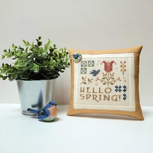 May include: A beige pillow with a white embroidered design featuring a red tulip, blue birds, and the text "HELLO SPRING!" The pillow is on a white surface with a small potted plant and a blue bird figurine.
