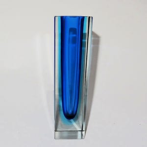 Könnte beinhalten: Eine rechteckige Glasvase mit leuchtend blauem Innenraum und klarer Außenseite. Die Vase hat ein schlankes, modernes Design mit einer langen, schmalen Öffnung. Die blaue Farbe ist im Zentrum konzentriert und erzeugt einen auffälligen visuellen Kontrast.